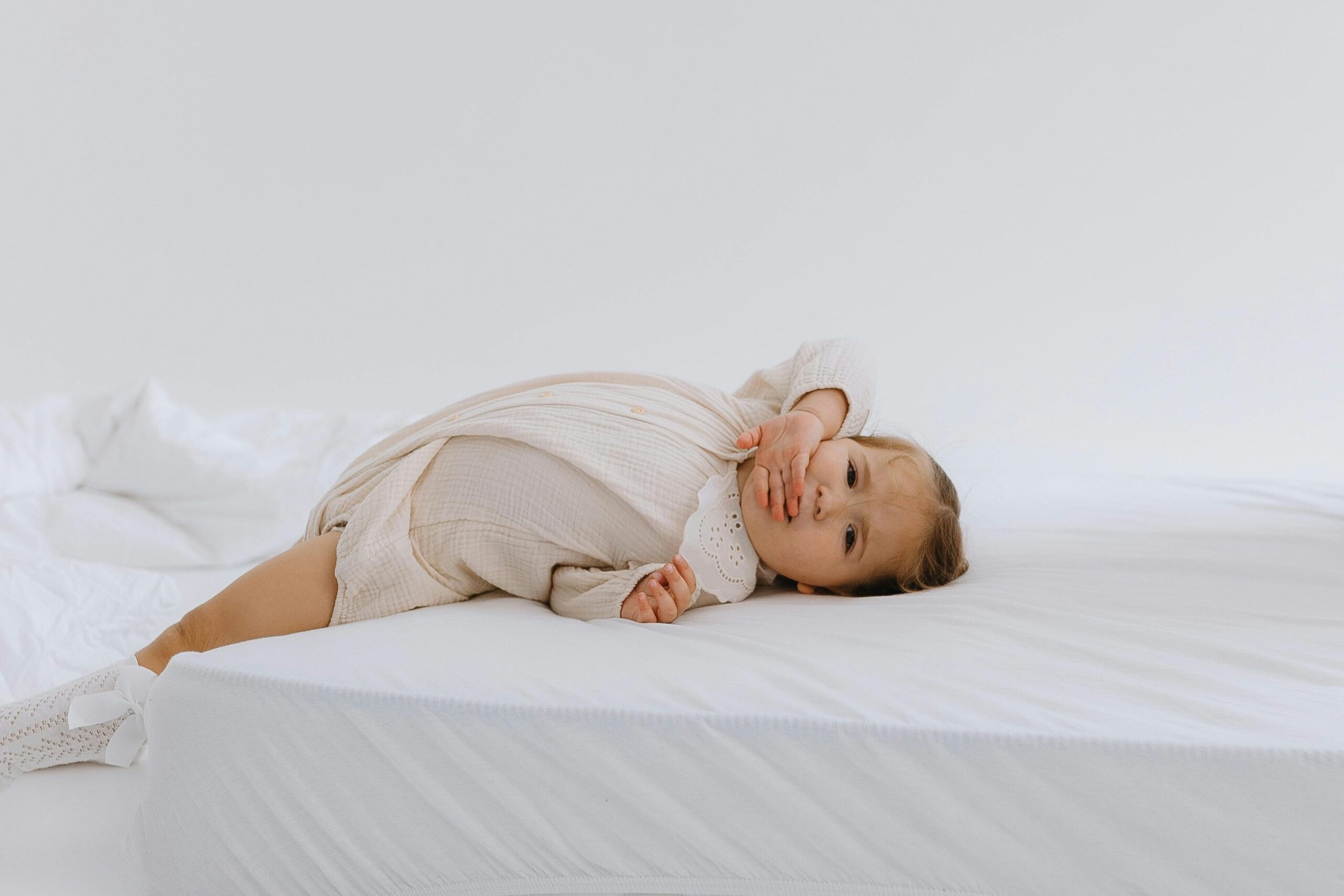 Cute baby in soft pajamas lying on a white mattress in a serene, minimalist bedroom setting.